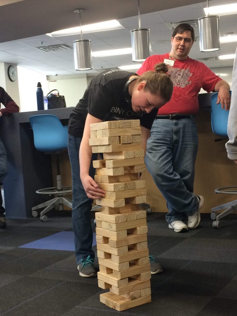 Four smiling people are watching one person place a block on a giant Jenga set. The person placing the block has blonde hair pulled into a low bun and is hunched over the Jenga game.