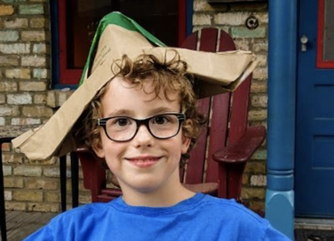A young boy, about 7 years old, is smiling at the camera. He is wearing black framed glasses, a blue t-shirt, and a makeshift boat hat made out of a paper bag. He has blonde curly hair.